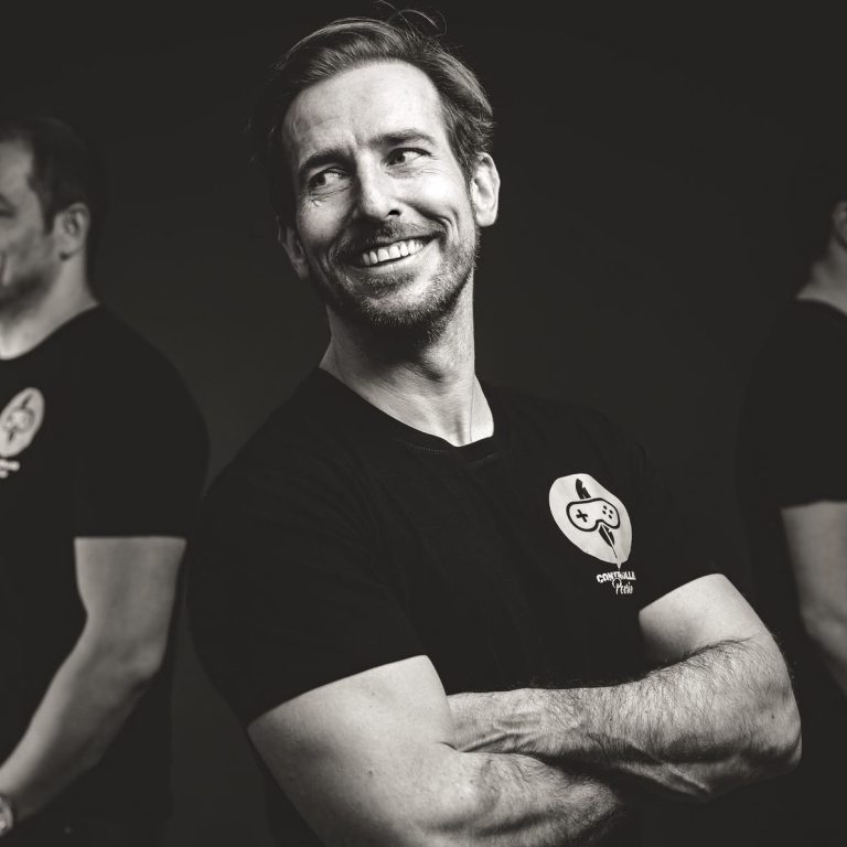 Controller Poesie – Studio-Portrait Raketenjansel Foto von Creator Raketenjansel in schwarzem T-Shirt mit Controller-Poesie-Logo, seitlich stehend und lächelnd vor dunklem Hintergrund.